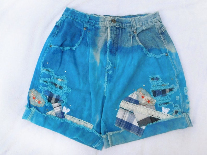 May include: Blue denim shorts with distressed details, patches, and embroidery. The shorts have a high waist and a relaxed fit.