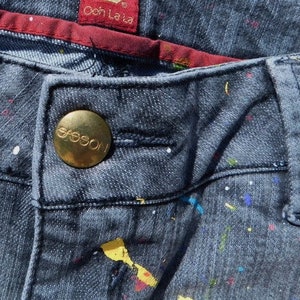 May include: Close-up of a pair of dark blue jeans with a gold button that says "Guess" on it. The jeans have a white and colorful paint splatter pattern.
