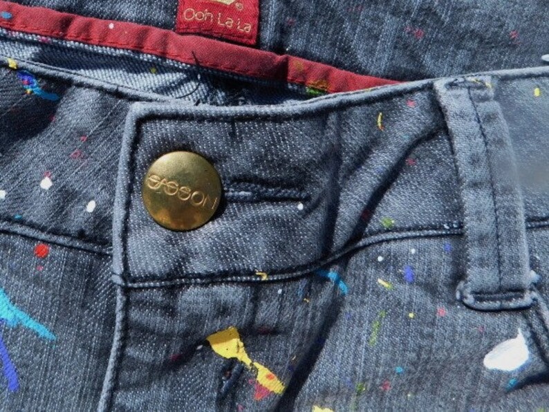 May include: Close-up of a pair of dark blue jeans with a gold button that says "Guess" on it. The jeans have a white and colorful paint splatter pattern.