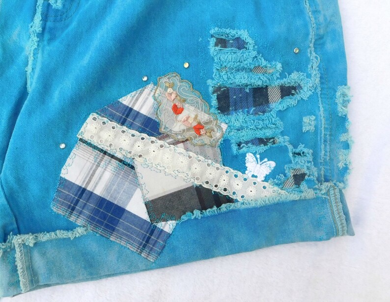 May include: A pair of blue denim shorts with patches of plaid fabric, lace, and a small embroidered design. The shorts have a distressed look with frayed edges.