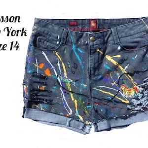 May include: Gray denim shorts with paint splatters in various colors, including white, yellow, orange, blue, and purple. The shorts have a distressed look with rips and frayed edges. The shorts have a small embroidered image of a cartoon horse on the right leg. The shorts have a label that reads "Sasson New York Size 14".
