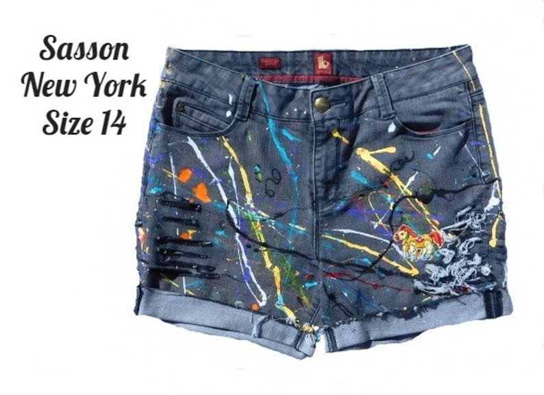 May include: Gray denim shorts with paint splatters in various colors, including white, yellow, orange, blue, and purple. The shorts have a distressed look with rips and frayed edges. The shorts have a small embroidered image of a cartoon horse on the right leg. The shorts have a label that reads "Sasson New York Size 14".