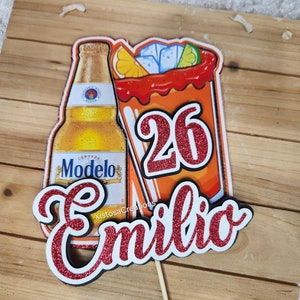 May include: A red and white cake topper with a beer bottle, a glass of red liquid, and the number 26. The name "Emilio" is written in red glitter.