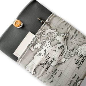 May include: A gray fabric notebook cover with a world map design on the front pocket. The cover is secured with a wooden button and a loop. A pen is tucked into the pocket, and a white card is visible inside.