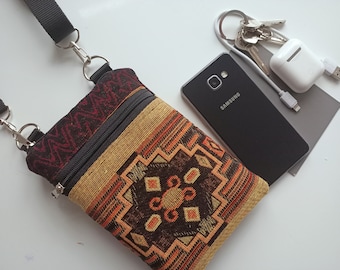 May include: A small, colorful, patterned crossbody bag with a zipper closure. The bag is made of fabric and has a black strap. The bag is lying on a white surface next to a black Samsung phone, a white charging case, and a set of keys.