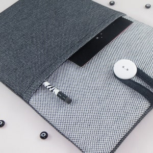 May include: A gray and white fabric tablet sleeve with a black elastic closure and a white button. A pencil is sticking out of the sleeve.