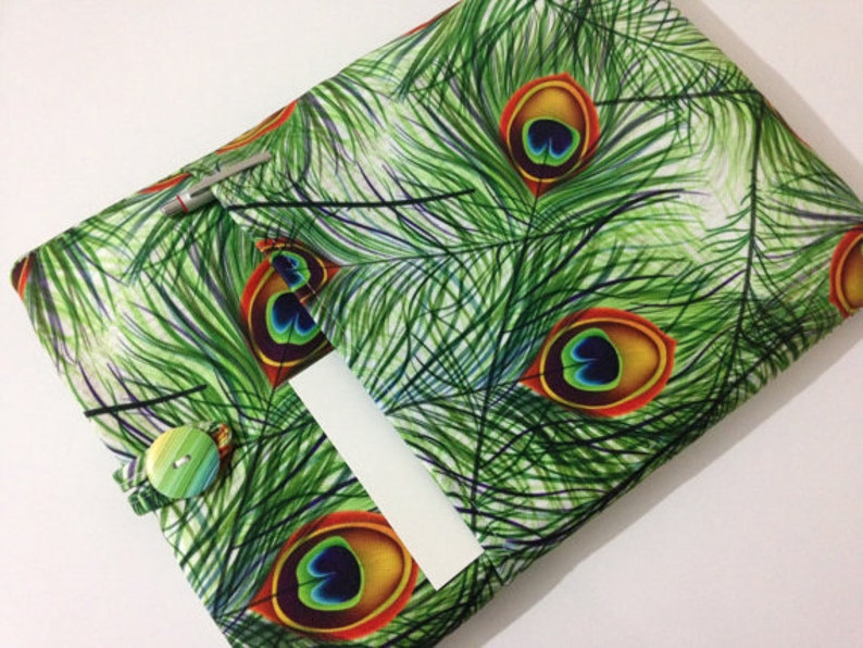 Pode incluir: Uma capa de livro de tecido verde com um padr&atilde;o de penas de pav&atilde;o. A capa tem um bolso na frente e um fecho de bot&atilde;o.