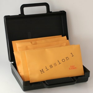 Peut inclure: Une mallette noire avec une poignée est ouverte et remplie de plusieurs enveloppes brunes. L'enveloppe du dessus porte l'inscription "Mission 1" en lettres d'imprimerie noires. Les mots "Top Secret" sont imprimés en rouge sur l'enveloppe.