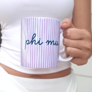 May include: A ceramic mug featuring a vertical lavender and white striped pattern. The text phi mu is printed in a dark blue cursive script across the centre of the mug. The mug has a white handle and a white interior rim.
