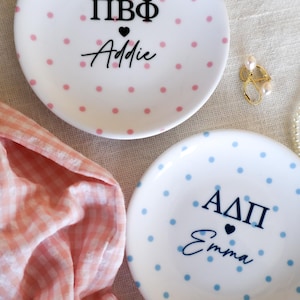 May include: Two round white ceramic trinket dishes with polka dots. One dish features pink dots with black text reading ΠΒΦ and Addie separated by a heart. The other dish features blue dots with dark blue text reading ΑΔΠ and Emma separated by a heart.
