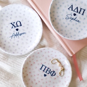 May include: Three small, white ceramic dishes with a floral pattern and dark blue Greek letters and names. One dish holds gold earrings with pearls. A pink notebook is in the background.
