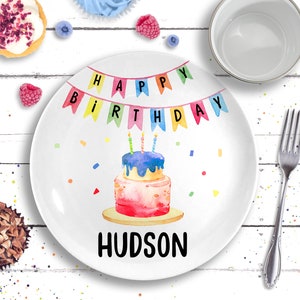 May include: White ceramic plate with a watercolor birthday cake design. The plate has the text "HAPPY BIRTHDAY" in colorful banner letters and the name "HUDSON" below the cake. The plate is surrounded by a fork, a mug, and some pastries.