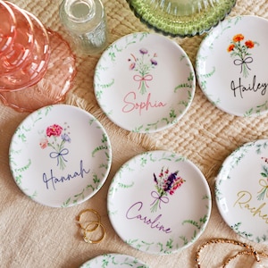 May include: Five white ceramic ring dishes with floral designs and personalized names: Sophia, Hailey, Hannah, Caroline, and Andrew. The dishes are on a beige textured surface.