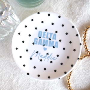 May include: A white ceramic trinket dish with black polka dots. The dish has the words "DELTA GAMMA" in light blue, with a small blue heart, and the name "Maddie" in cursive. A gold beaded bracelet is next to the dish.