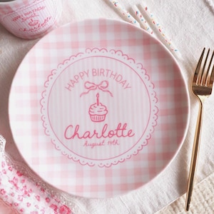 May include: A round ceramic plate and matching mug featuring a pink and white gingham pattern. The plate displays the text Happy Birthday, a cupcake illustration with a bow, the name Charlotte, and the date August 14th inside a scalloped border.
