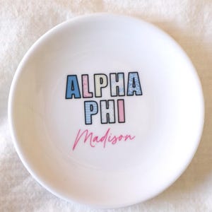 May include: A white ceramic trinket dish with the words "ALPHA PHI" in blue, pink, and light blue block letters, and the name "Madison" in pink script. The dish is round and sits on a white surface.