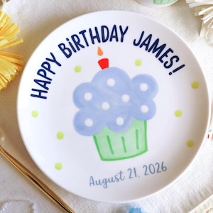 May include: A white ceramic plate featuring a hand-painted blue cupcake with white polka dots, a green base, and a single lit candle. The text reads HAPPY BIRTHDAY JAMES! and August 21, 2026 in blue lettering, surrounded by small yellow dots.