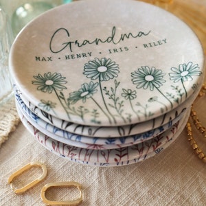 May include: A white ceramic trinket dish with a floral design and the text "Grandma Max. Henry. Iris. Riley" in black script. The dish is stacked on top of other similar dishes with different floral designs.