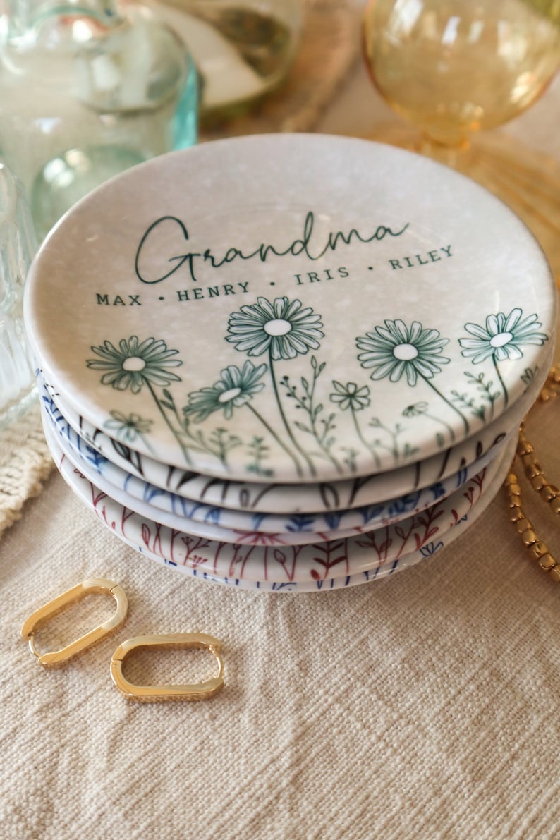 May include: A white ceramic trinket dish with a floral design and the text "Grandma Max. Henry. Iris. Riley" in black script. The dish is stacked on top of other similar dishes with different floral designs.