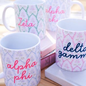 May include: Four white ceramic mugs featuring a repeating scalloped pattern with floral motifs. Each mug displays a Greek organization name in cursive script: Zeta in pink, alpha chi in pink, alpha phi in pink, and delta gamma in dark blue.