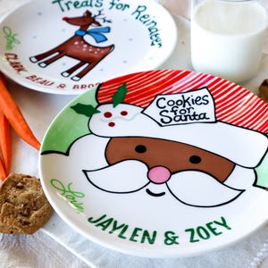 May include: Two white ceramic plates with Christmas-themed designs. One plate features a reindeer with the text "Treats for Reindeer" and the other plate features Santa Claus with the text "Cookies for Santa". Both plates have green and red accents and are personalized with the names "Jaylen & Zoey".