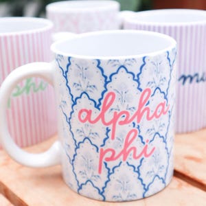 May include: A white ceramic mug featuring a blue and white scalloped floral pattern with the text alpha phi written in a pink cursive font. The mug has a curved handle and is positioned in front of other mugs with striped patterns.