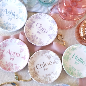 May include: A collection of six small round ceramic trinket dishes featuring floral patterns and personalized names including Ashley, Clara, Anna, Chloe, Hallie, and Sadie, each accented with a small bow icon and descriptive text.