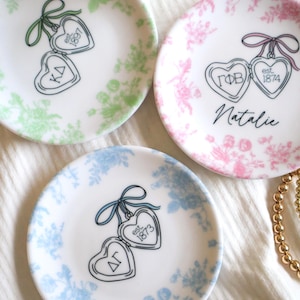 May include: Three small, white decorative plates with floral patterns in green, pink, and blue. Each plate features a heart-shaped pendant design with Greek letters and the year 1873 or 1874. One plate has the name "Natalie".