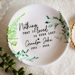 May include: A white ceramic trinket dish with a green botanical design and the text "Nothing that is loved is ever lost. Grandpa John 1941 - 2026." The dish is surrounded by greenery and a gold beaded bracelet.
