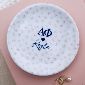 May include: A white ceramic trinket dish with a floral pattern of small pink roses. The dish has the text "AΦ" above a heart and the name "Kayla" in navy blue. A pair of gold earrings with pearl drops rests nearby.
