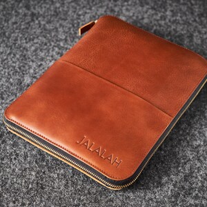 Peut inclure: Une pochette en cuir marron avec une fermeture éclair dorée et le mot "JALALAH" en relief sur le devant. La pochette a une poche sur le devant. La pochette rectangulaire est probablement pour une tablette ou un ordinateur portable.