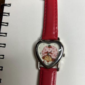 K&ouml;nnte beinhalten: Eine Armbanduhr mit rotem Lederarmband und herzf&ouml;rmigem Zifferblatt. Das Zifferblatt zeigt eine Zeichentrickfigur mit einem rosa Erdbeerhut. Die Uhr hat ein silberfarbenes Metallgeh&auml;use.