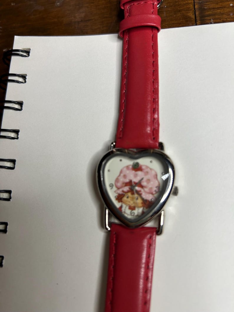 K&ouml;nnte beinhalten: Eine Armbanduhr mit rotem Lederarmband und herzf&ouml;rmigem Zifferblatt. Das Zifferblatt zeigt eine Zeichentrickfigur mit einem rosa Erdbeerhut. Die Uhr hat ein silberfarbenes Metallgeh&auml;use.
