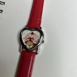 K&ouml;nnte beinhalten: Eine Armbanduhr mit rotem Lederarmband und herzf&ouml;rmigem Zifferblatt. Das Zifferblatt zeigt eine Zeichentrickfigur mit einem rosa Erdbeerhut. Die Uhr hat eine silberfarbene L&uuml;nette und ein rotes Lederarmband.