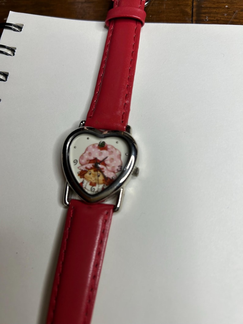 K&ouml;nnte beinhalten: Eine Armbanduhr mit rotem Lederarmband und herzf&ouml;rmigem Zifferblatt. Das Zifferblatt zeigt eine Zeichentrickfigur mit einem rosa Erdbeerhut. Die Uhr hat eine silberfarbene L&uuml;nette und ein rotes Lederarmband.
