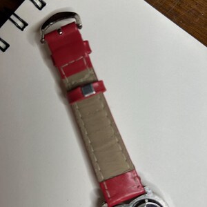 K&ouml;nnte beinhalten: Ein rotes Uhrenarmband mit einer silbernen Schnalle und einem herzf&ouml;rmigen Zifferblatt. Auf dem Zifferblatt steht "Strawberry Shortcake". Das Armband besteht aus rotem Leder mit einer beigefarbenen Unterseite.