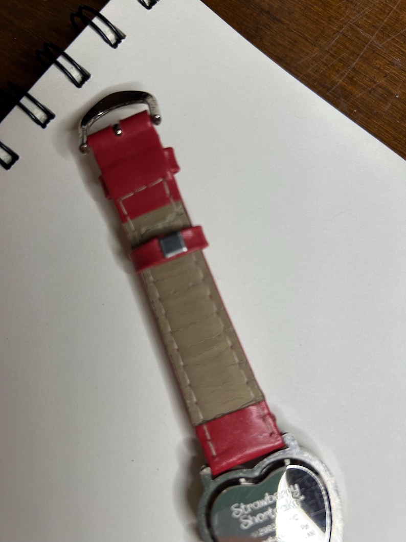 K&ouml;nnte beinhalten: Ein rotes Uhrenarmband mit einer silbernen Schnalle und einem herzf&ouml;rmigen Zifferblatt. Auf dem Zifferblatt steht "Strawberry Shortcake". Das Armband besteht aus rotem Leder mit einer beigefarbenen Unterseite.