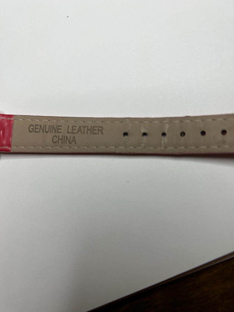 K&ouml;nnte beinhalten: Nahaufnahme eines roten Uhrenarmbands mit den Worten "GENUINE LEATHER CHINA" auf der Unterseite. Das Armband hat mehrere L&ouml;cher zur Anpassung.