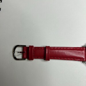 K&ouml;nnte beinhalten: Nahaufnahme eines roten Leder-Uhrenarmbands mit einer silbernen Schnalle. Das Armband ist an den R&auml;ndern gen&auml;ht und hat eine Schlaufe zur Sicherung der &uuml;bersch&uuml;ssigen L&auml;nge. Das Uhrenarmband befindet sich auf einer wei&szlig;en Oberfl&auml;che.