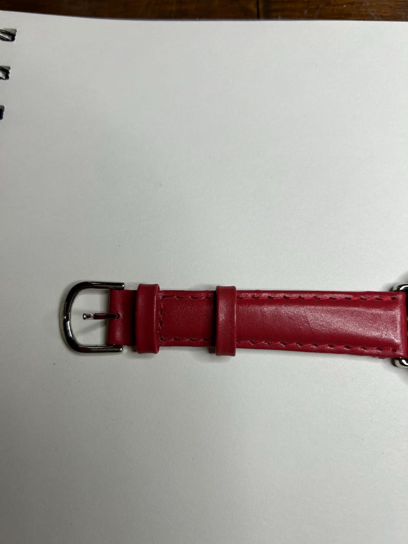 K&ouml;nnte beinhalten: Nahaufnahme eines roten Leder-Uhrenarmbands mit einer silbernen Schnalle. Das Armband ist an den R&auml;ndern gen&auml;ht und hat eine Schlaufe zur Sicherung der &uuml;bersch&uuml;ssigen L&auml;nge. Das Uhrenarmband befindet sich auf einer wei&szlig;en Oberfl&auml;che.