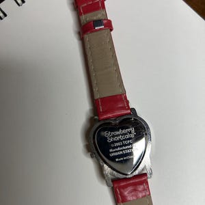 K&ouml;nnte beinhalten: Eine Strawberry Shortcake-Uhr mit herzf&ouml;rmigem Zifferblatt und rotem Lederarmband. Das Zifferblatt ist silberfarben mit dem Text "Strawberry Shortcake" und anderem Text. Das Armband ist rot und beige.