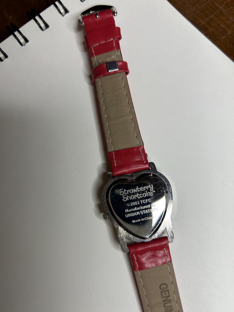 K&ouml;nnte beinhalten: Eine Strawberry Shortcake-Uhr mit herzf&ouml;rmigem Zifferblatt und rotem Lederarmband. Das Zifferblatt ist silberfarben mit dem Text "Strawberry Shortcake" und anderem Text. Das Armband ist rot und beige.