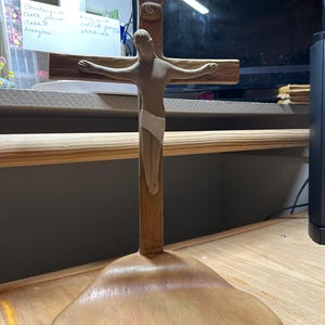 May include: A wooden crucifix with a figure of Jesus. The cross is made of light brown wood, with the letters INRI at the top. The figure is light-colored, with arms outstretched. The base is a rounded shape.