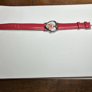 K&ouml;nnte beinhalten: Eine rote Armbanduhr mit herzf&ouml;rmigem Zifferblatt. Das Zifferblatt zeigt eine Zeichentrickfigur. Die Uhr hat ein rotes Lederarmband. Die Uhr liegt auf einer wei&szlig;en Oberfl&auml;che.