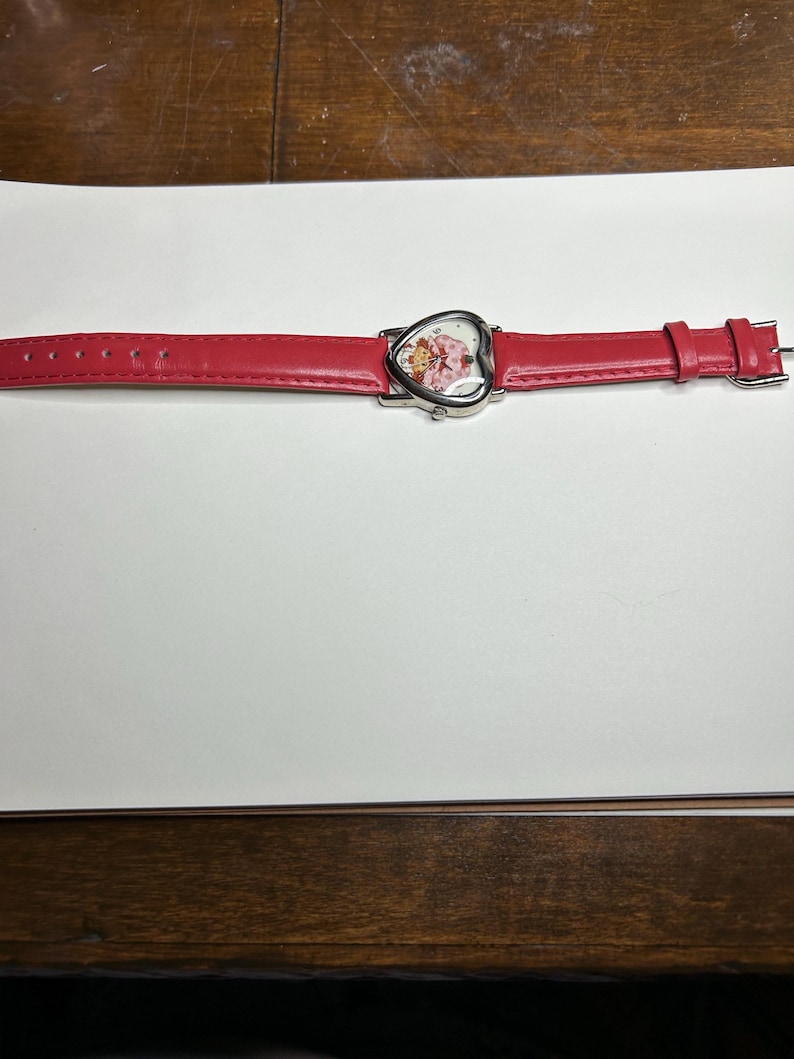 K&ouml;nnte beinhalten: Eine rote Armbanduhr mit herzf&ouml;rmigem Zifferblatt. Das Zifferblatt zeigt eine Zeichentrickfigur. Die Uhr hat ein rotes Lederarmband. Die Uhr liegt auf einer wei&szlig;en Oberfl&auml;che.