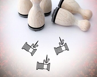 May include: Five wooden stamps with black ink pads, shaped like a rounded rectangle with a rounded top. The stamps are arranged on a white surface. Three stamps are standing upright, and two are lying down. The stamps are all the same size and shape. The stamps are blank, but there are three small drawings of spools of thread on the surface below the stamps.