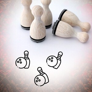 Könnte beinhalten: Satz mit fünf Holz-Bowling-Pinstempeln mit schwarzen Stempelkissen. Die Stempel sind auf einer weißen Oberfläche angeordnet. Drei Stempel stehen aufrecht, zwei liegen flach. Unter den Stempeln befinden sich drei Schwarzweiß-Zeichnungen von Bowling-Pins und -Kugeln.