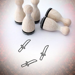 Könnte beinhalten: Fünf Holzstempel mit schwarzen Stempelflächen, die die Form eines abgerundeten Rechtecks haben. Die Stempel sind in einem Halbkreis auf einer weißen Oberfläche angeordnet. Vier kleine schwarz-weiße Messerillustrationen sind auf der Oberfläche unterhalb der Stempel gezeichnet.