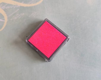 May include: A square, clear plastic ink pad with a vibrant pink ink surface. The ink pad is set against a light green background with decorative script and text. Ideal for crafting and stamping projects.