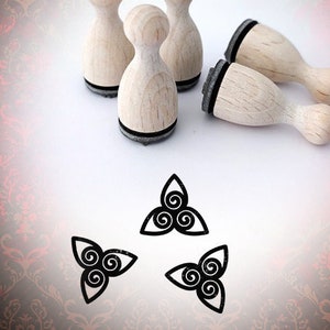 Könnte beinhalten: Vier Holzstempel mit schwarzen Stempelflächen und einem schwarz-weißen Design einer dreifachen Spirale. Die Stempel sind in einem Halbkreis auf einer weißen Oberfläche angeordnet. Der Hintergrund ist ein rotes und braunes Damastmuster mit dem Text "Bloody Nose" in Weiß.